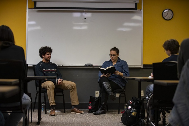 An image of a person reading from a book in front of a room of students